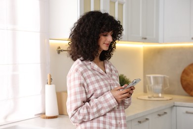 Beautiful young woman in stylish pyjama with smartphone in kitchen