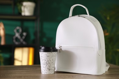 Stylish white backpack and cup of coffee on wooden table indoors, space for text