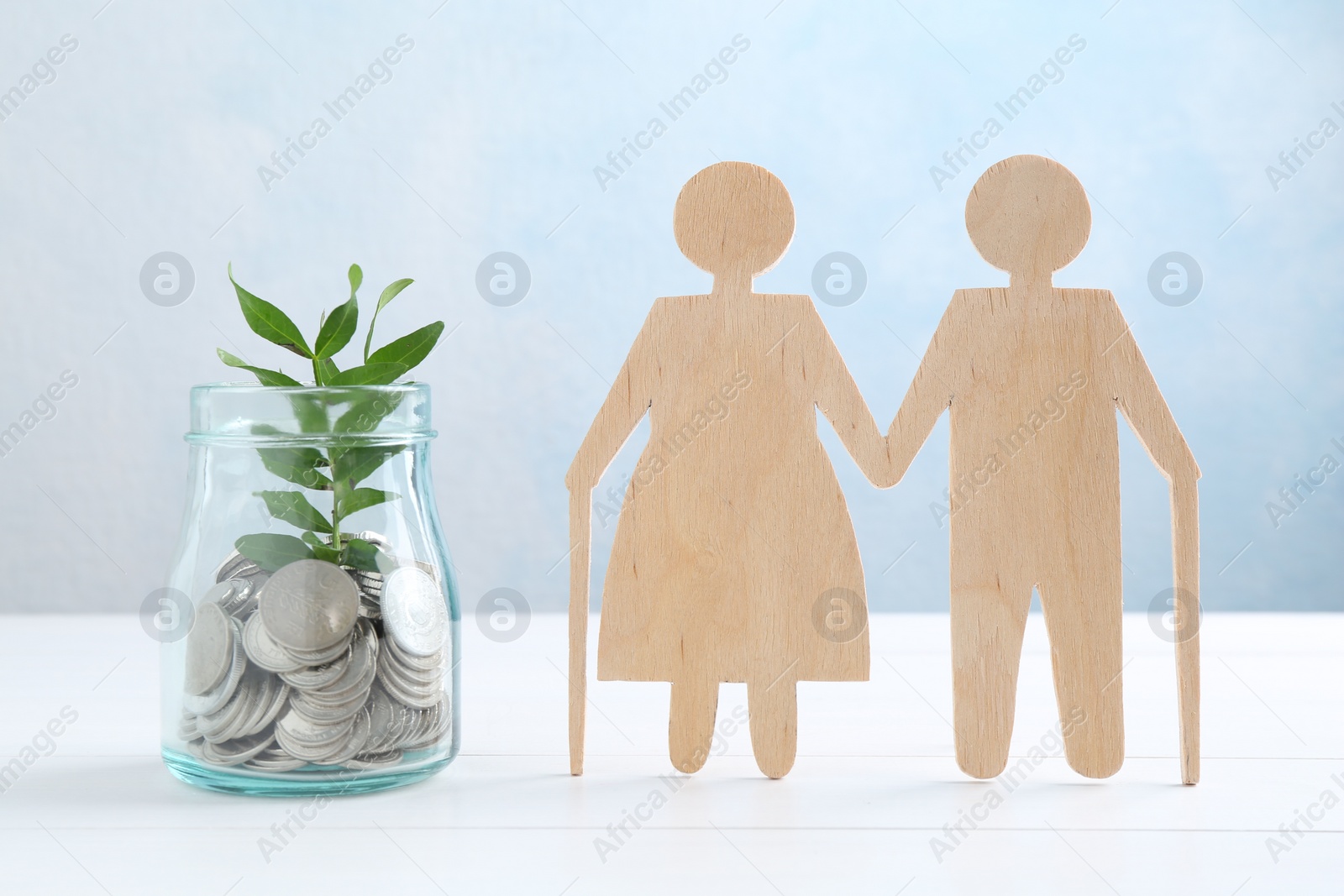 Photo of Pension savings. Figure of senior couple, jar of coins and green twig on white wooden table