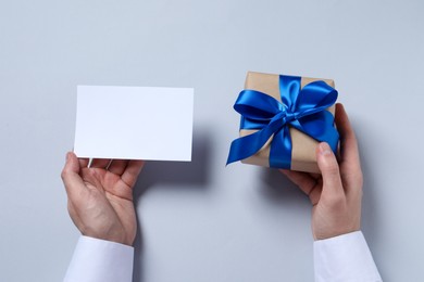 Photo of Man holding gift box with blue bow and blank greeting card on white background, top view