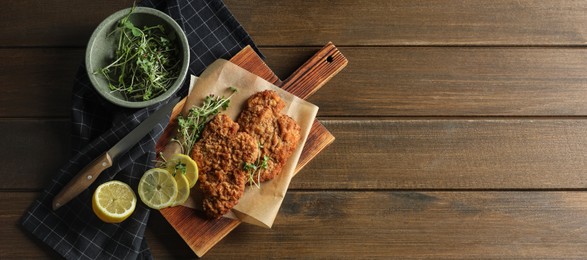 Tasty schnitzels served with lemon and microgreens on wooden table, flat lay. Banner design with space for text