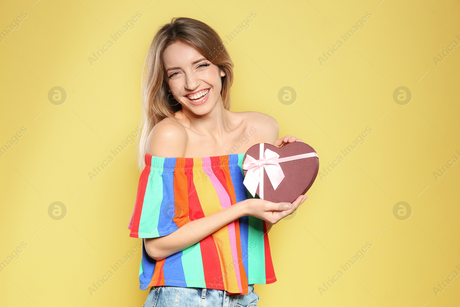 Photo of Portrait of beautiful smiling girl with heart shaped gift box on yellow background. International Women's Day