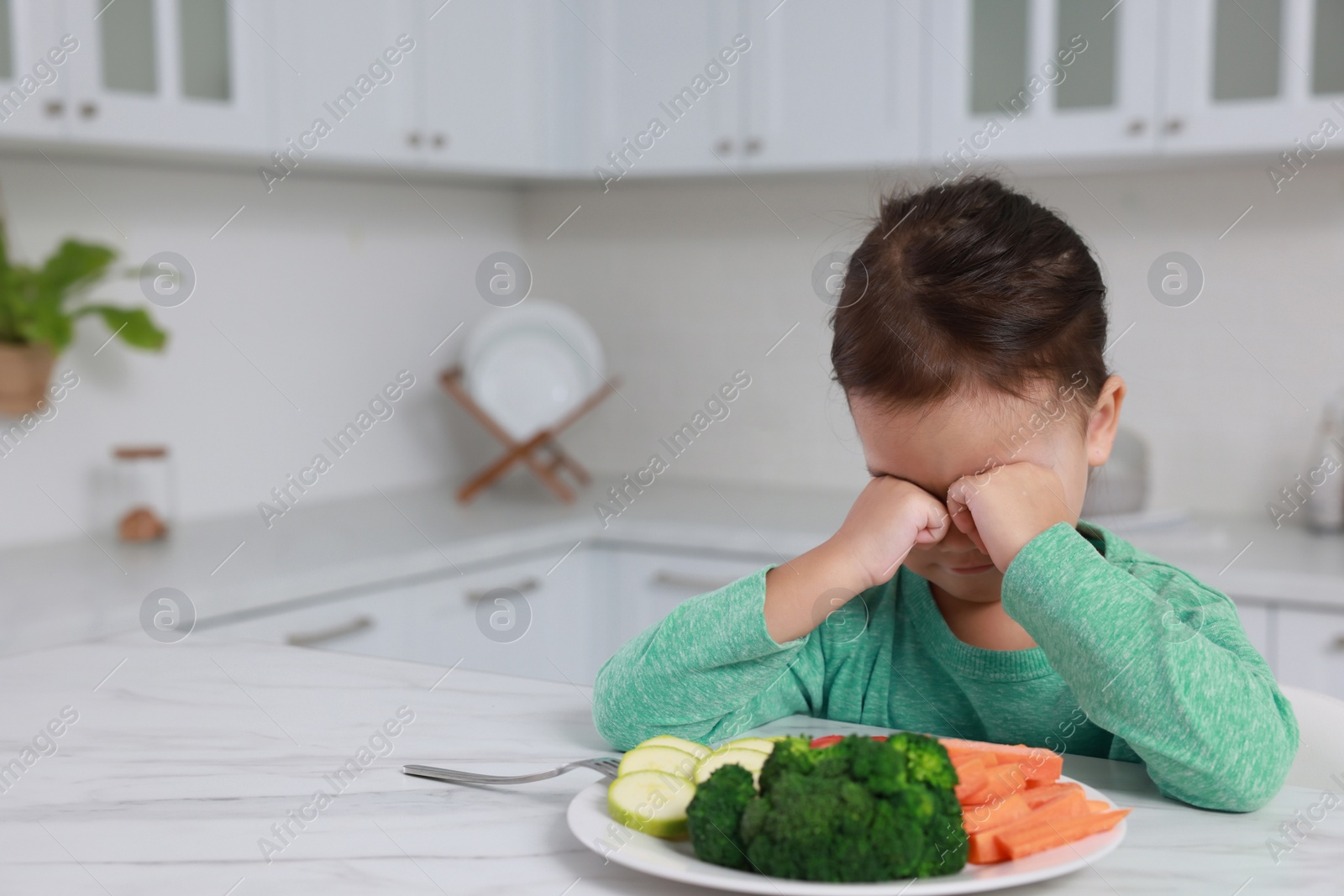 Photo of Cute little girl crying and refusing to eat vegetables in kitchen, space for text