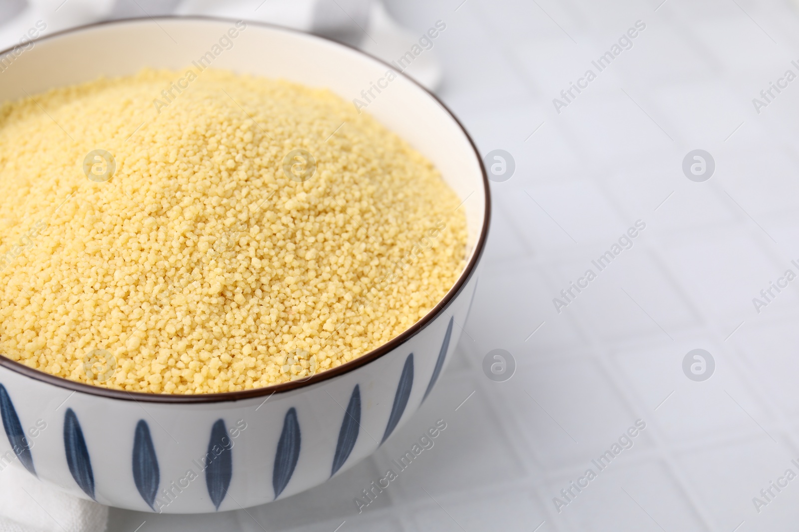 Photo of Raw couscous in bowl on white tiled table, closeup. Space for text