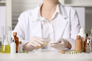 Photo of Dermatologist with bottle testing cosmetic product at white table indoors, closeup