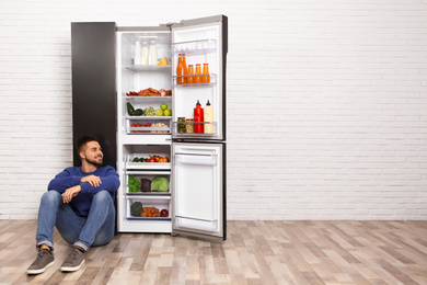 Photo of Happy young man sitting on floor near refrigerator indoors, space for text