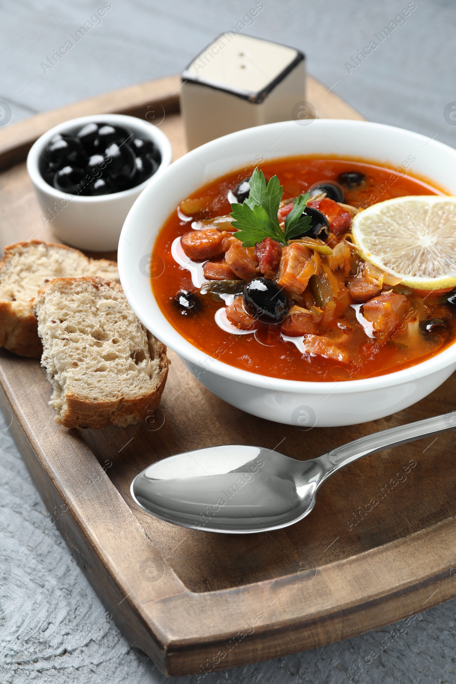 Photo of Meat solyanka soup with sausages, olives and vegetables in bowl served on grey wooden table, closeup