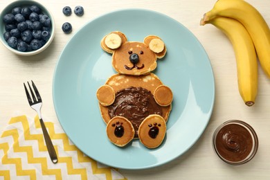 Photo of Creative serving for kids. Plate with cute bear made of pancakes, blueberries, bananas and chocolate paste on light wooden table, flat lay