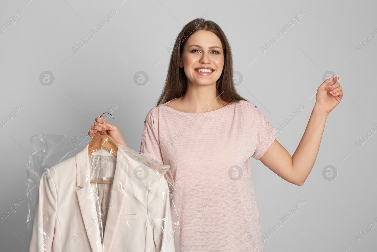 Photo of Young woman holding hanger with jacket in plastic bag on light grey background. Dry-cleaning service