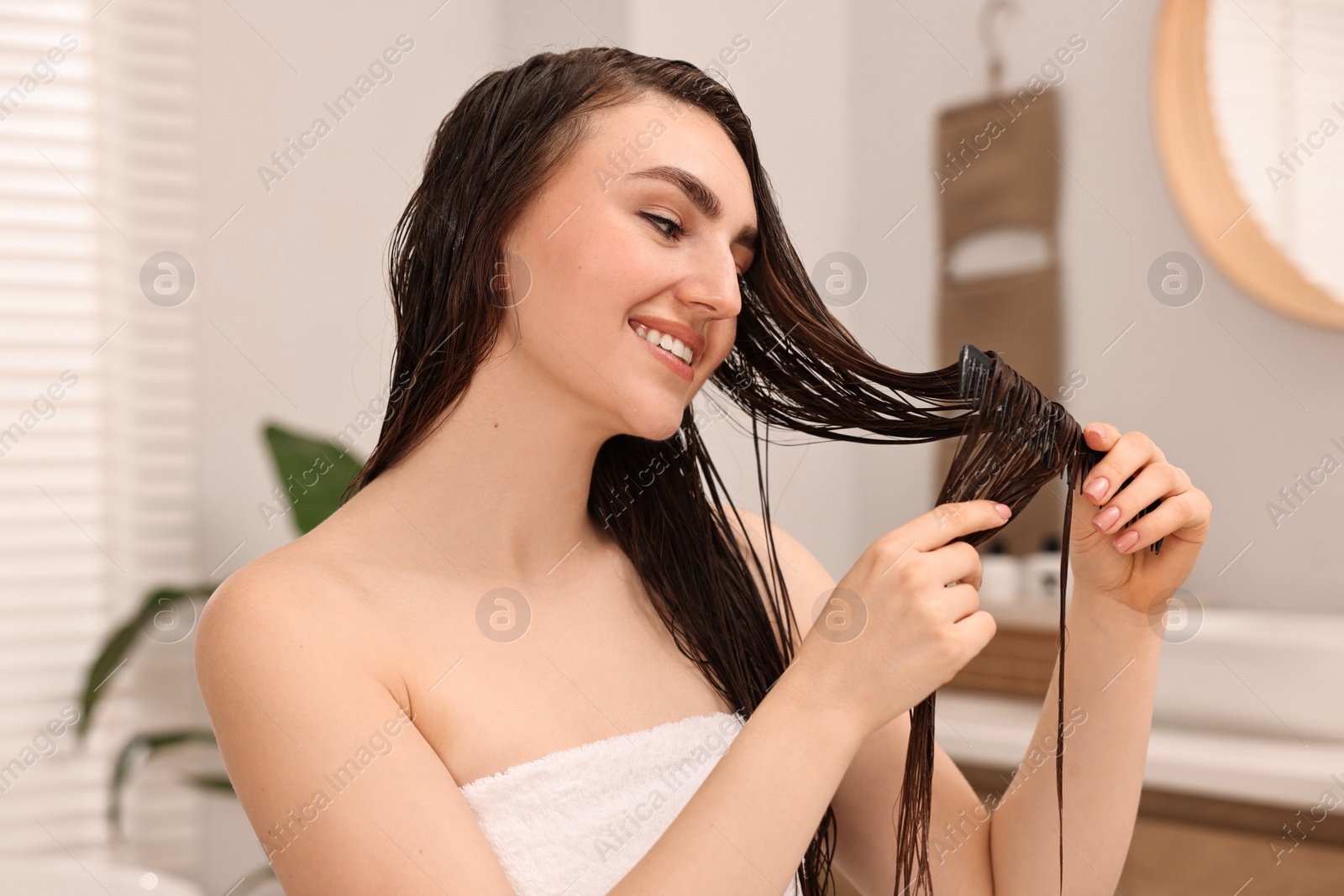 Photo of Young woman brushing hair after applying mask in bathroom