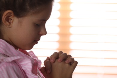Photo of Cute little girl with hands clasped together praying near window, closeup. Space for text