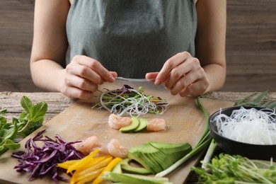 Woman wrapping spring roll at wooden table with products, closeup