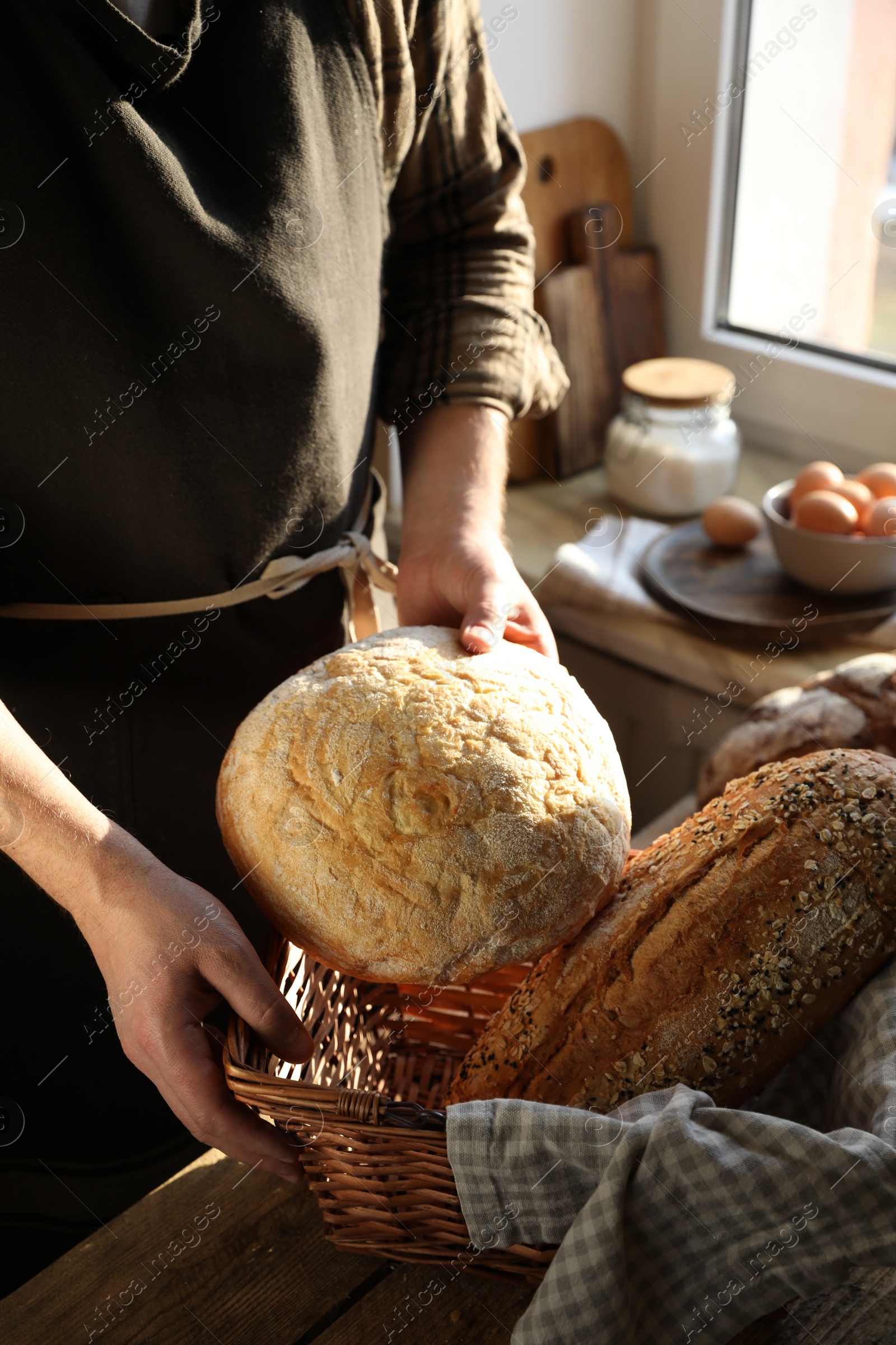 Photo of Man holding wicker basket with different types of bread at wooden table indoors, closeup