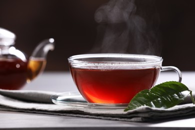 Fresh hot tea in glass cup with saucer, teapot and leaf on table, closeup