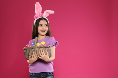 Little girl in bunny ears headband holding basket with Easter eggs on color background, space for text