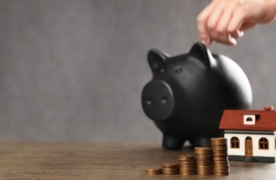 Photo of Savings for house purchase. Woman putting coin into piggy bank at wooden table, focus on money. Space for text