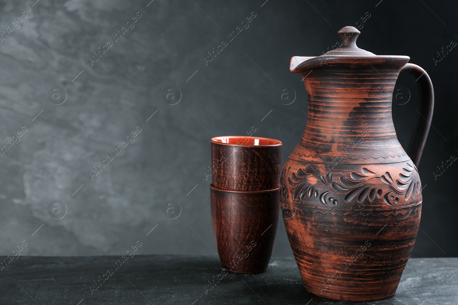 Photo of Brown clay jug and cups on black table, space for text. Handmade utensils