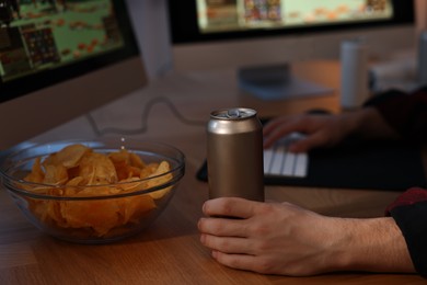 Young man with energy drink and chips playing video game at wooden desk indoors, closeup