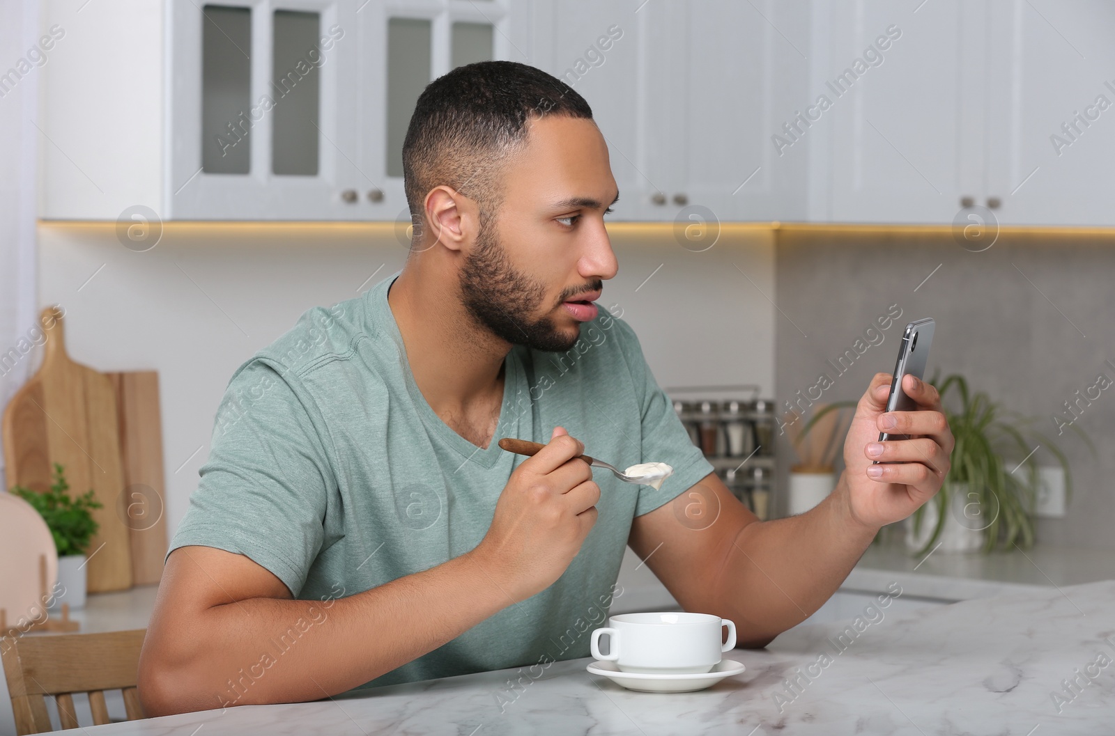 Photo of Young man using smartphone while having breakfast at white marble table in kitchen. Internet addiction