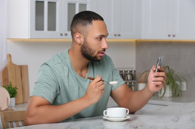 Young man using smartphone while having breakfast at white marble table in kitchen. Internet addiction