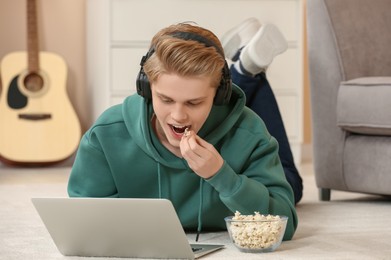 Photo of Teenage boy with headphones eating popcorn while using laptop at home