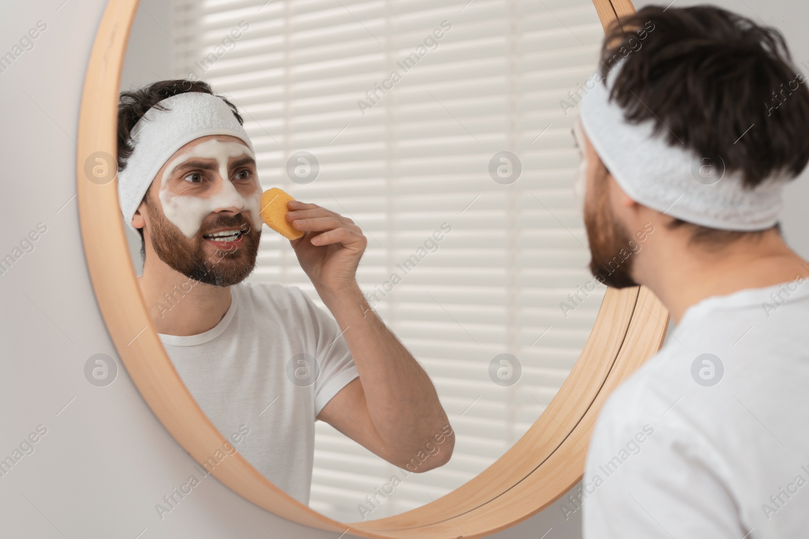 Photo of Man with headband washing his face using sponge near mirror in bathroom