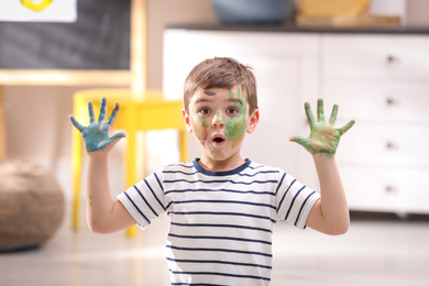 Emotional little child with paint on face and palms in room