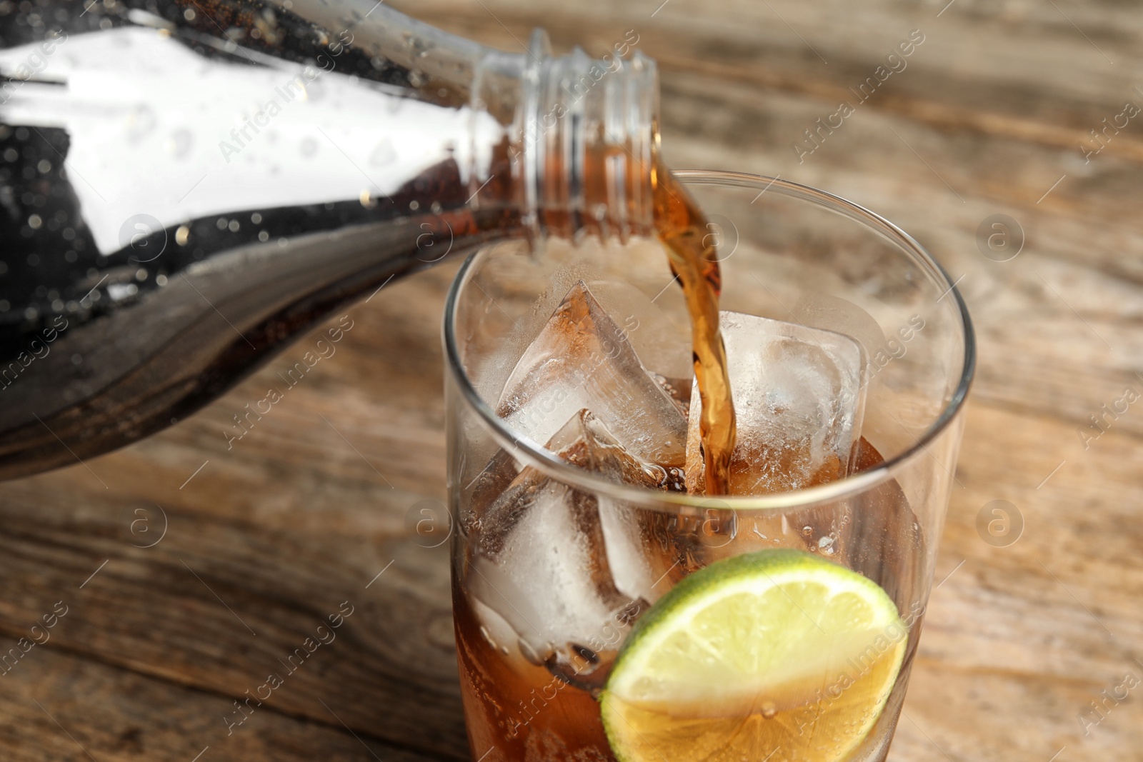 Photo of Pouring refreshing soda drink into glass with ice cubes and lime on wooden table, closeup