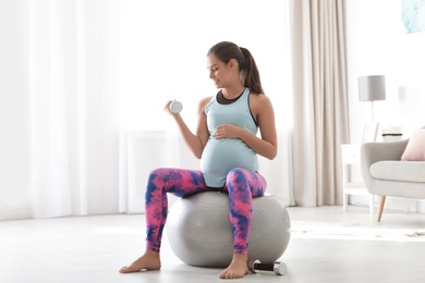 Photo of Young pregnant woman in fitness clothes lifting dumbbell at home