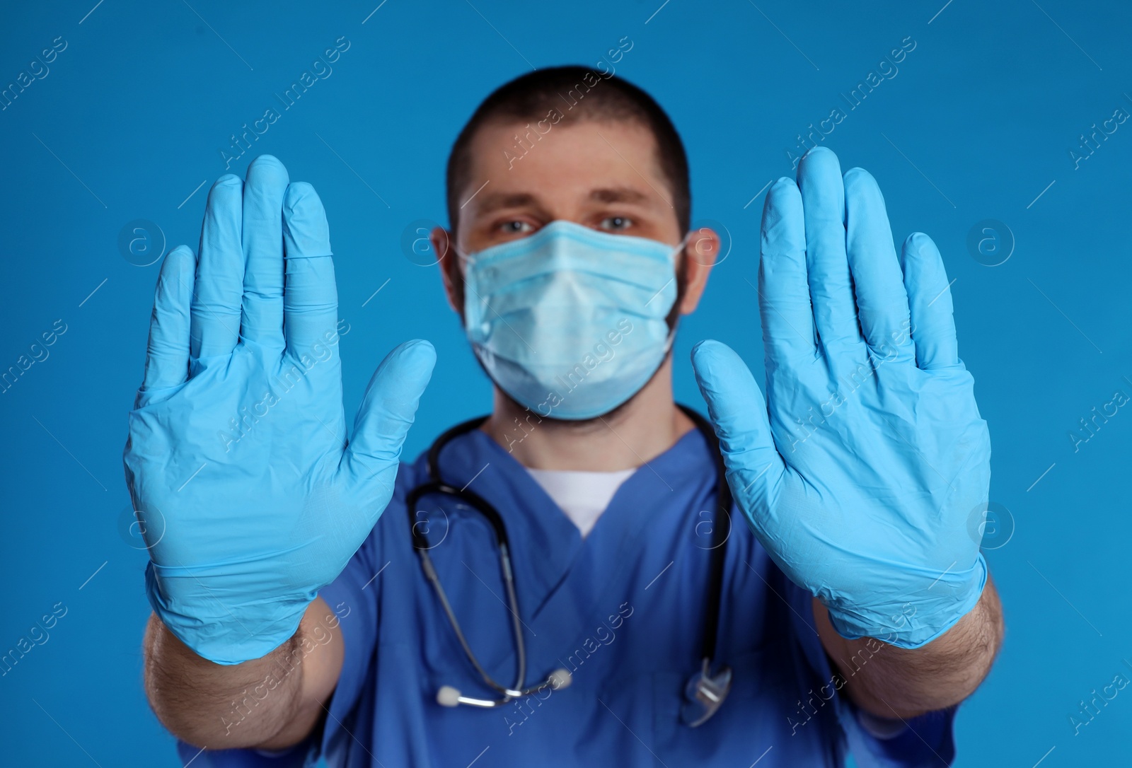 Photo of Doctor in protective mask showing stop gesture on light blue background. Prevent spreading of coronavirus