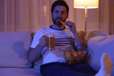 Photo of Man eating chips and drinking beer while watching TV on sofa at night. Bad habit