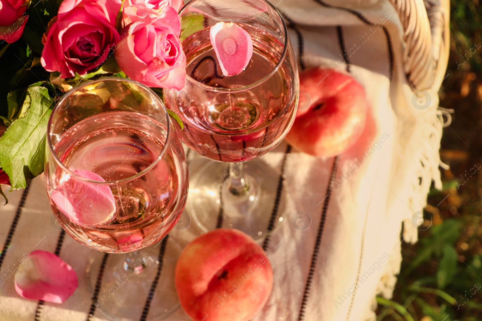 Photo of Glasses of delicious rose wine with petals, flowers and peaches on white picnic blanket outside, above view