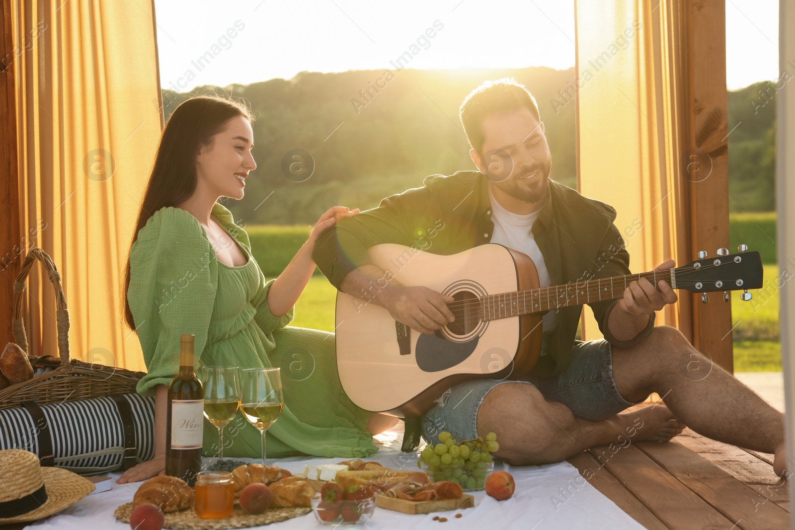 Photo of Romantic date. Beautiful woman listening to her boyfriend playing guitar during picnic outdoors