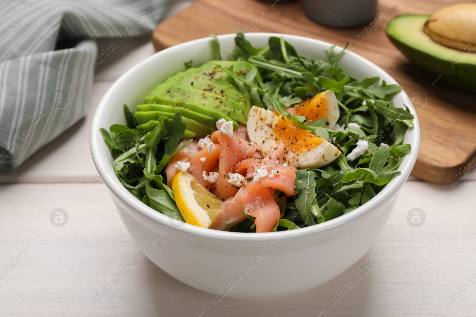 Photo of Delicious salad with boiled egg, salmon and avocado in bowl on white wooden table, closeup