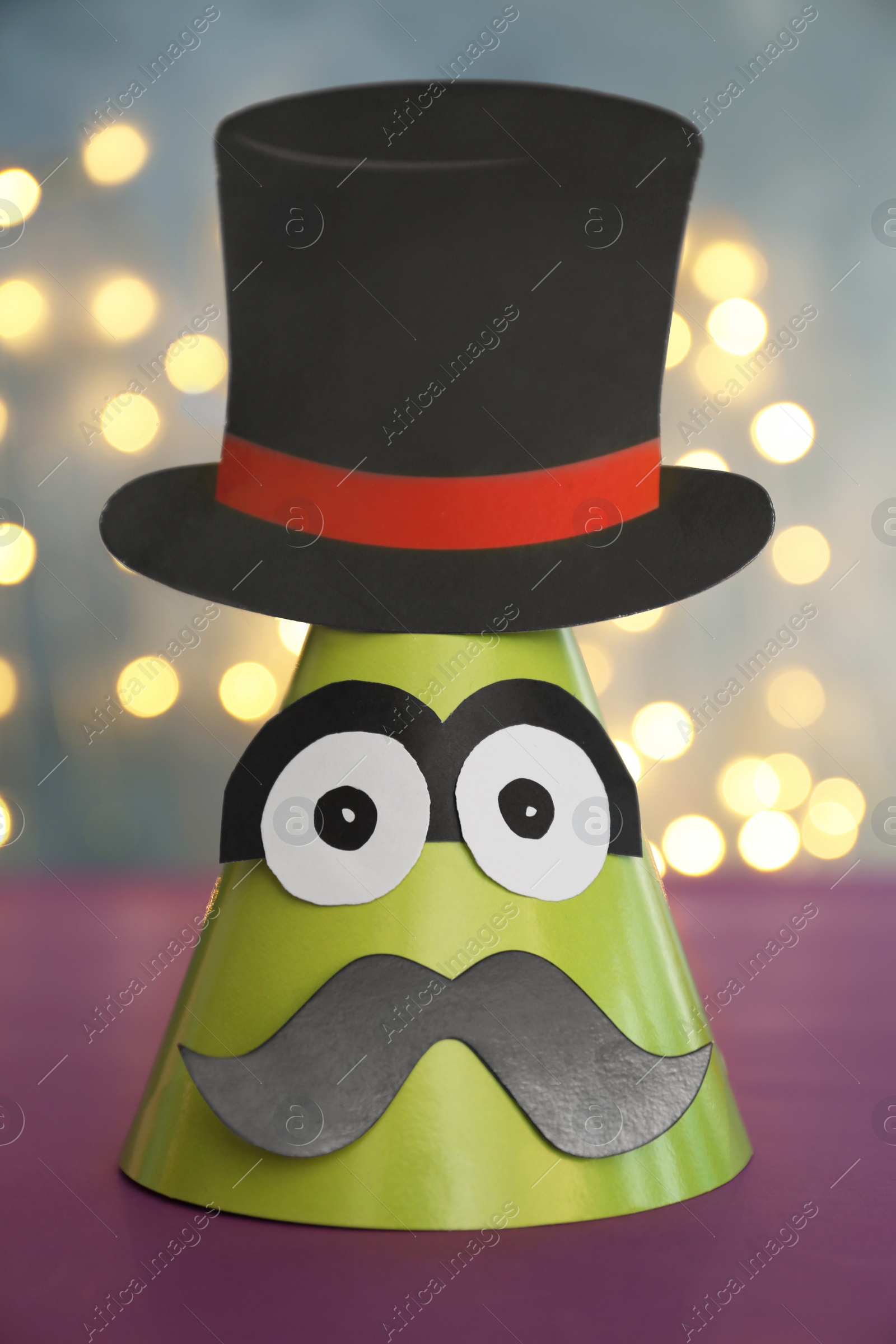 Photo of Funny handmade party hat on purple table against blurred lights, closeup