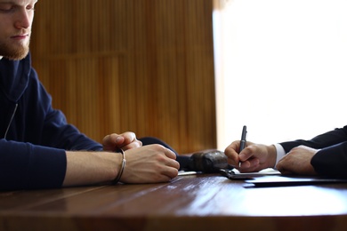 Photo of Police officer interrogating criminal in handcuffs at desk indoors