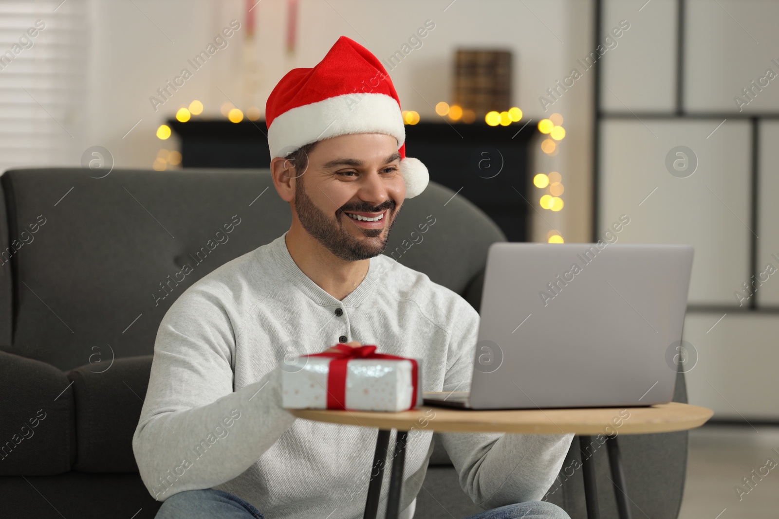 Photo of Celebrating Christmas online with exchanged by mail presents. Happy man in Santa hat with gift box during video call on laptop at home