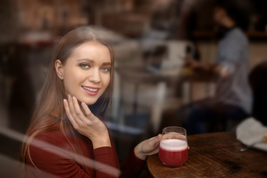 Pretty young woman with cocktail at table in cafe, view from outdoors through window