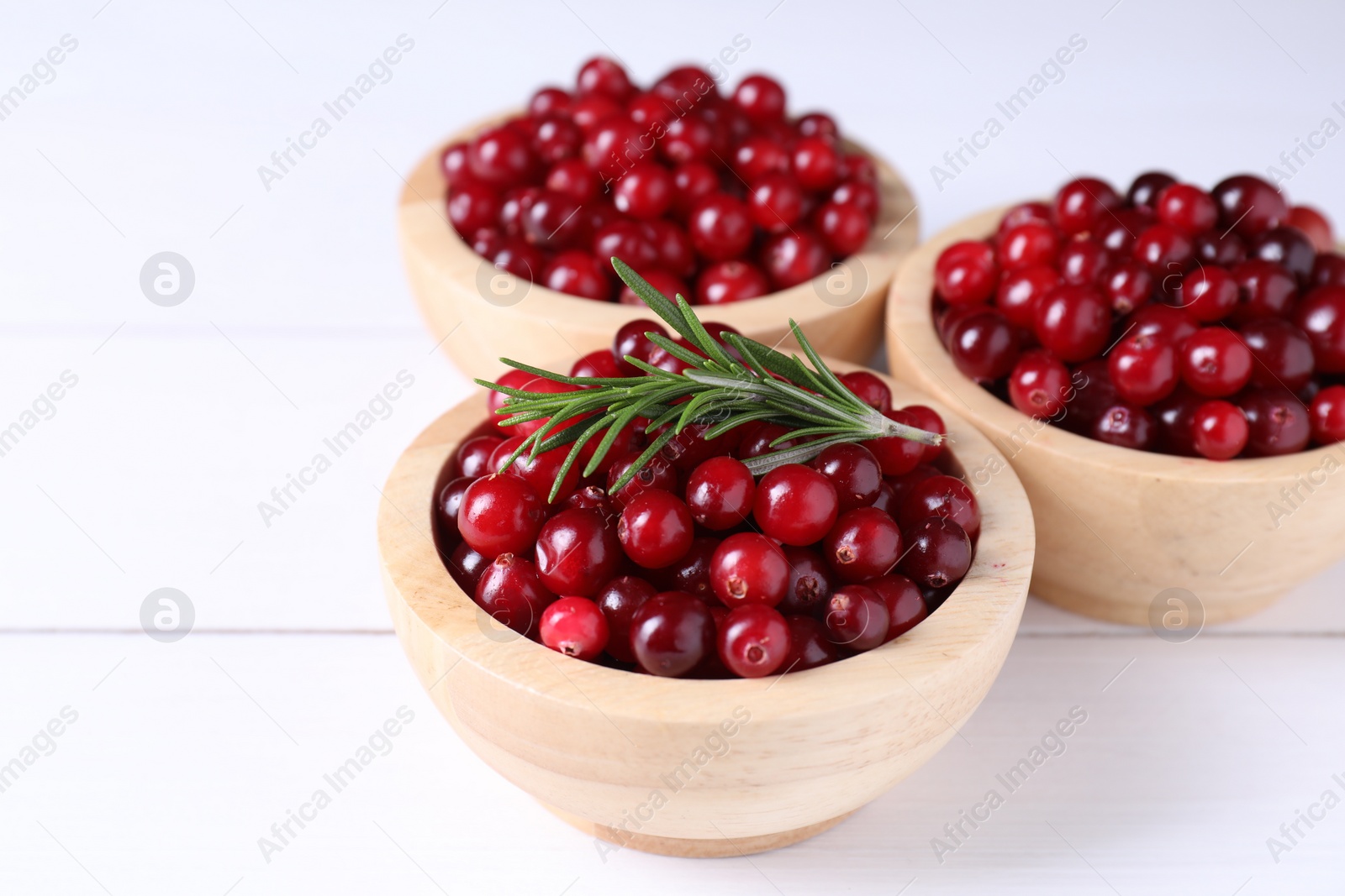 Photo of Fresh ripe cranberries in bowls and rosemary on white wooden table, closeup