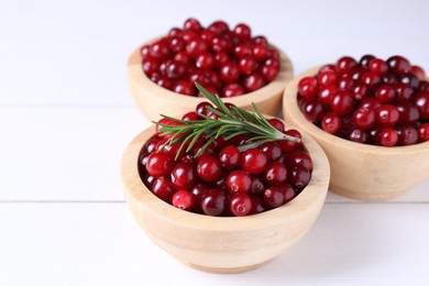 Photo of Fresh ripe cranberries in bowls and rosemary on white wooden table, closeup