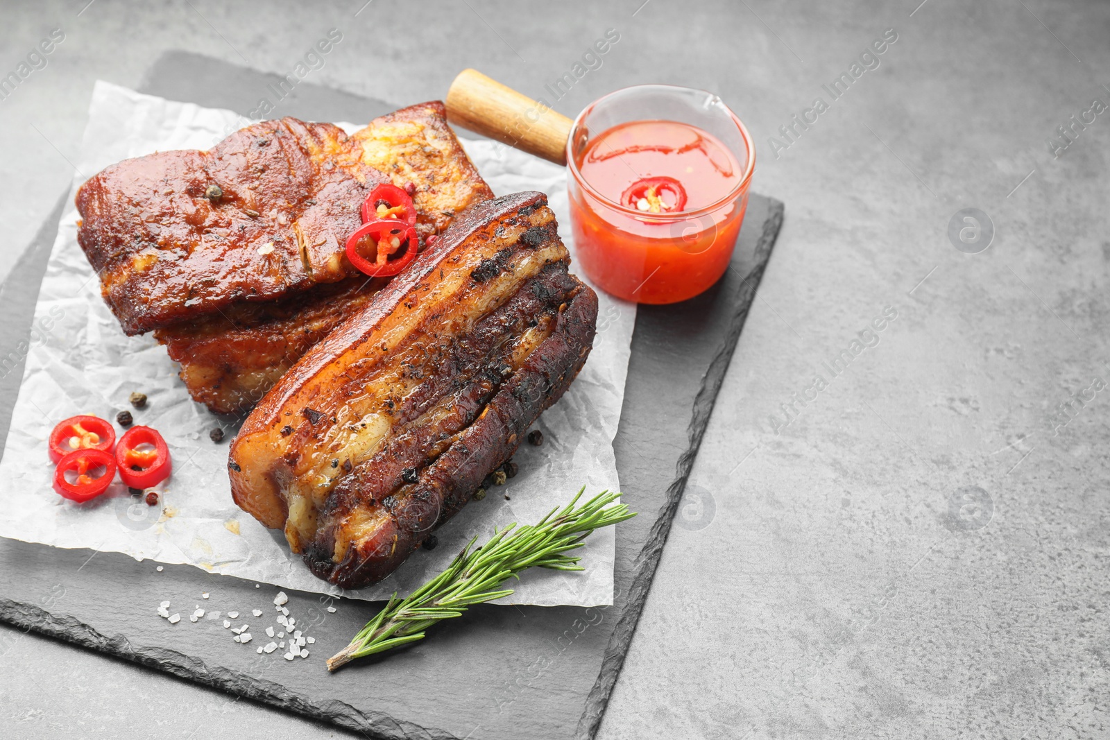 Photo of Pieces of baked pork belly served with sauce, rosemary and chili pepper on grey table, space for text