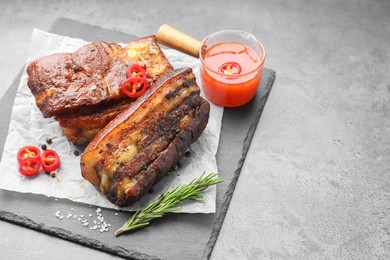 Photo of Pieces of baked pork belly served with sauce, rosemary and chili pepper on grey table, space for text