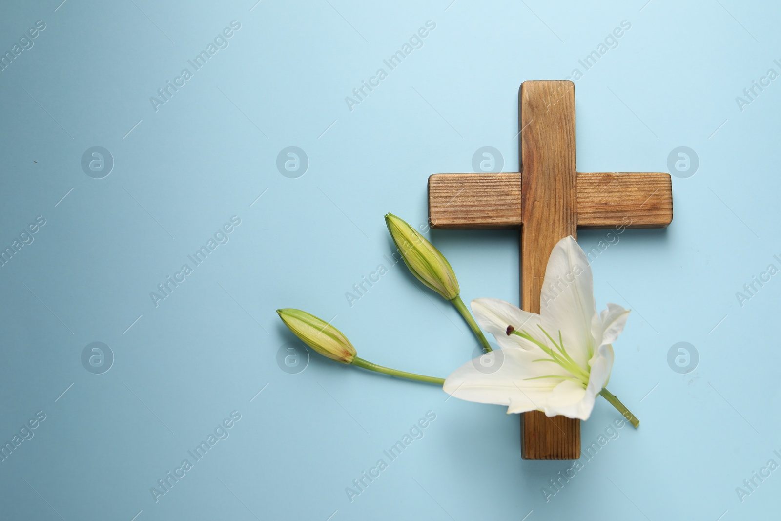 Photo of Wooden cross and lily flowers on light blue background, top view with space for text. Easter attributes