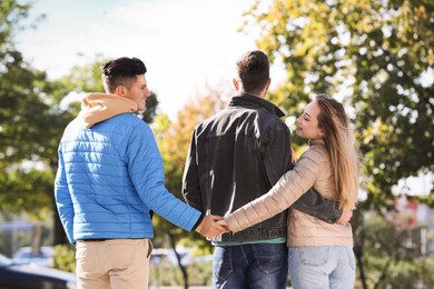 Photo of Woman holding hands with another man behind her boyfriend's back during walk in park. Love triangle