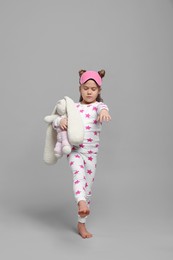 Girl in pajamas and sleep mask with toy bunny sleepwalking on gray background