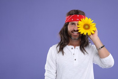 Photo of Stylish hippie man covering eye with sunflower on violet background, space for text