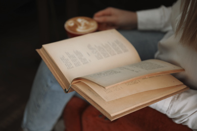 Woman with cup of coffee reading book indoors, closeup