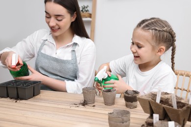 Mother with her daughter spraying water onto vegetable seeds in pots at wooden table indoors