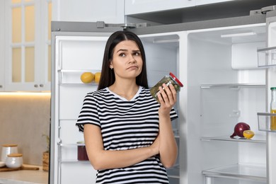 Upset woman with jar of pickles near empty refrigerator in kitchen