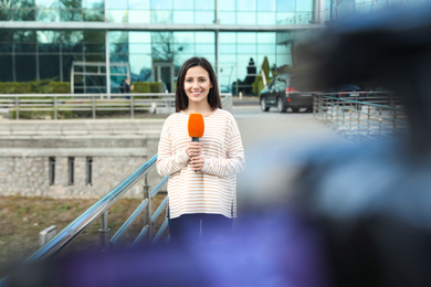 Photo of Young female journalist with microphone working on city street